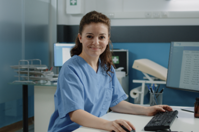 a white woman in light blue scrubs smiles at the camera while standing at a computer desk